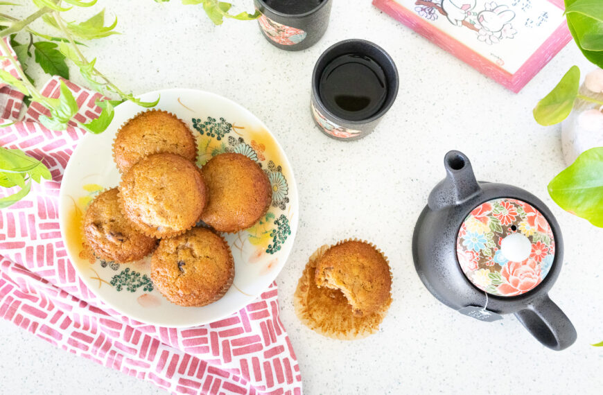 A flatlay photo of fuyu persimmon muffins on a colorful Japanese floral plate. Beneath the plate is a rose pink and white tea cloth. To the right of the plate is a muffin with a bite taken out of it and a Japanese tea pot and cup. There are also some green plants and a photo of bunnies poking into the frame.