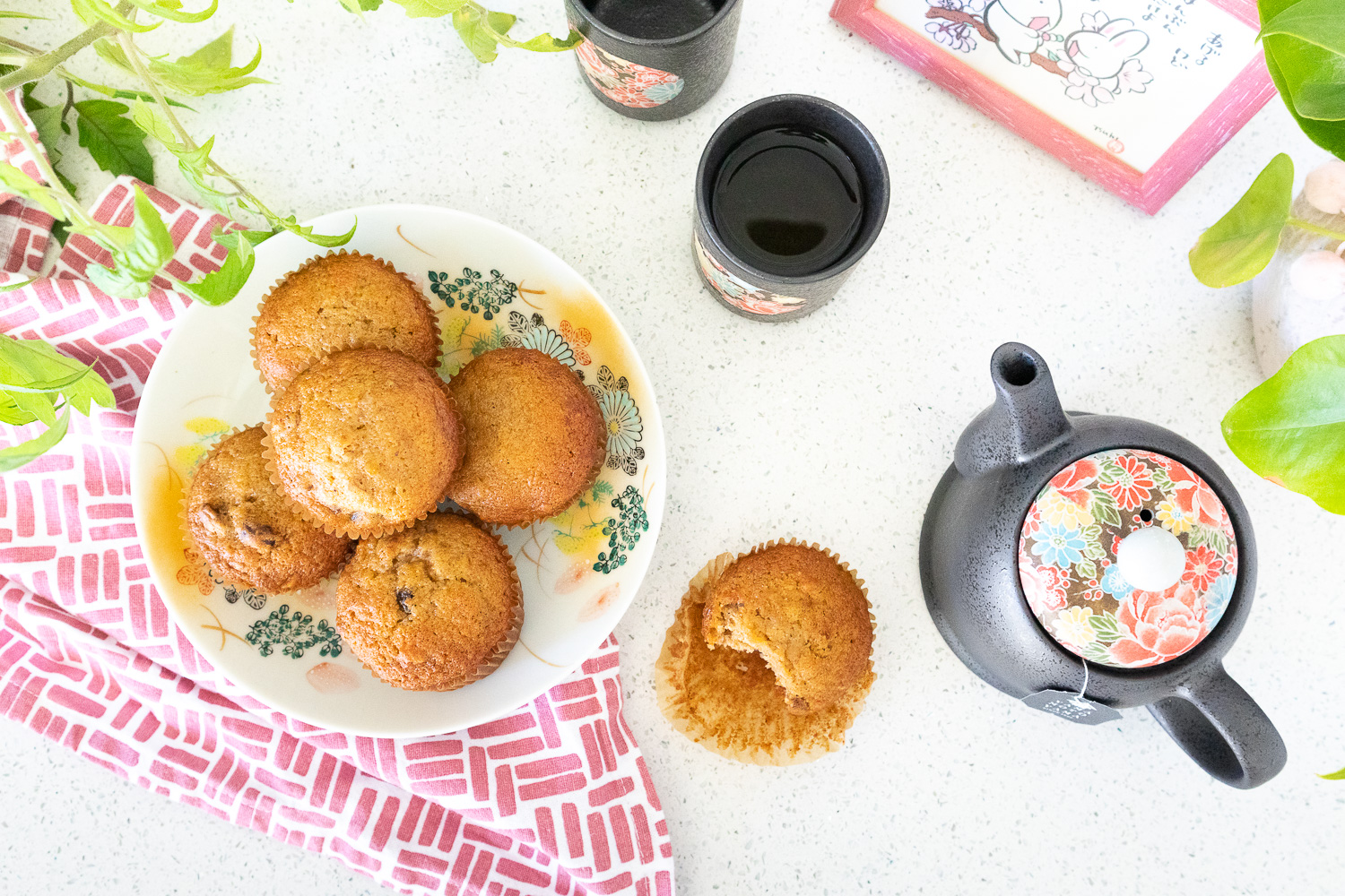 A flatlay photo of fuyu persimmon muffins on a colorful Japanese floral plate. Beneath the plate is a rose pink and white tea cloth. To the right of the plate is a muffin with a bite taken out of it and a Japanese tea pot and cup. There are also some green plants and a photo of bunnies poking into the frame.