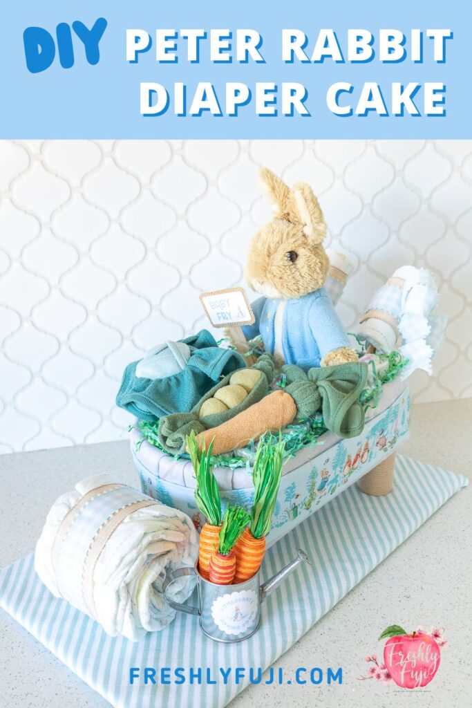 A photo for Pinterest featuring a Peter Rabbit wheelbarrow diaper cake. A tan plush rabbit with a light blue jacket is sitting in a diaper cake made out of baby blankets and diapers. Sitting on top of the wheelbarrow are vegetables (cabbage, pea pods, carrots) made from washcloths. The wheelbarrow wheel and handles are also made using diapers. 