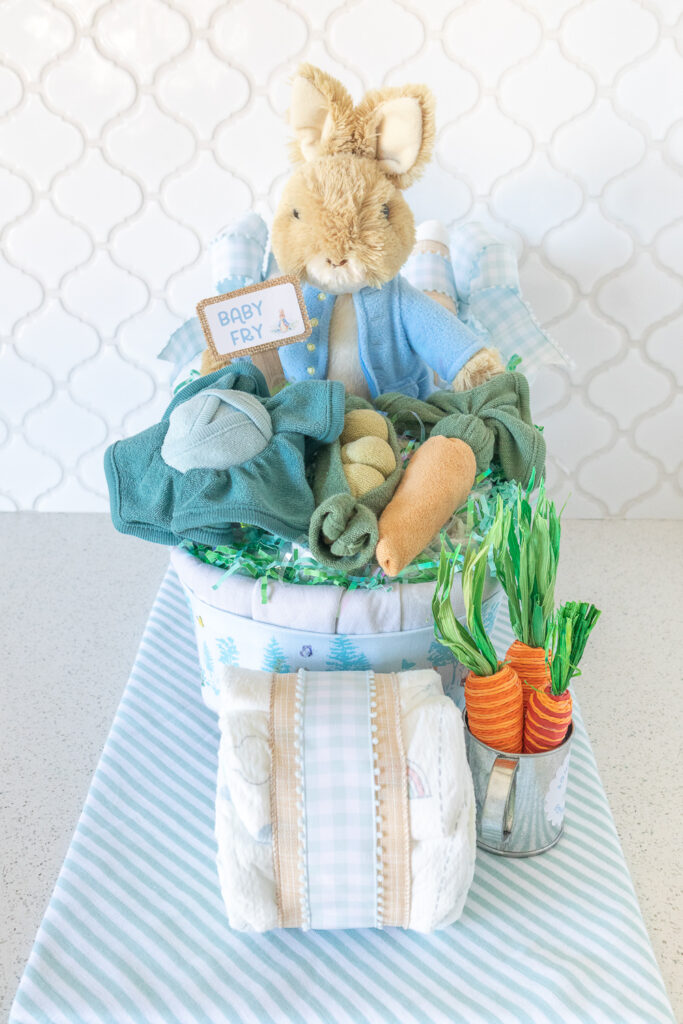 A photo of the front of a Peter Rabbit wheelbarrow diaper cake. A tan plush rabbit with a light blue jacket is sitting in a diaper cake made out of baby blankets and diapers. Sitting on top of the wheelbarrow are vegetables (cabbage, pea pods, carrots) made from washcloths. 