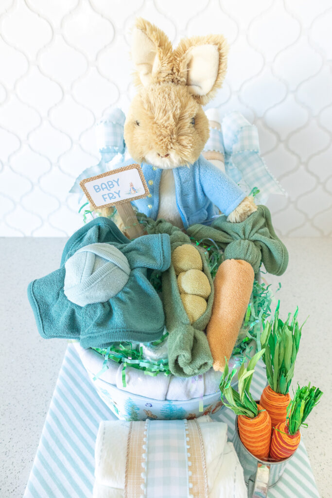 A closeup photo of a Peter Rabbit wheelbarrow diaper cake. A tan plush rabbit with a light blue jacket is sitting in a diaper cake made out of baby blankets and diapers. Sitting on top of the wheelbarrow are vegetables (cabbage, pea pods, carrots) made from washcloths. 