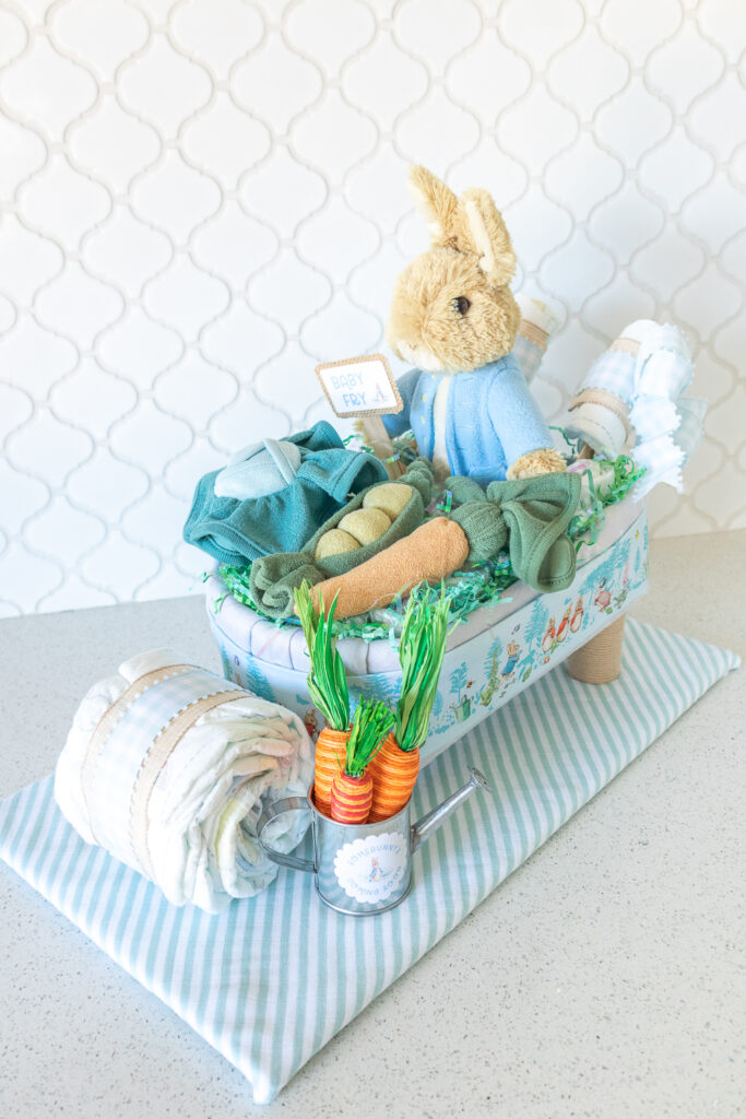 A photo of a handmade Peter Rabbit wheelbarrow diaper cake on an angle. A tan plush rabbit with a light blue jacket is sitting in a diaper cake made out of baby blankets and diaper. Sitting on top of the wheelbarrow are vegetables (cabbage, pea pods, carrots) made from washcloths. The wheelbarrow is wrapped in a strip of blue Peter Rabbit fabric. The wheel and handles are made from diapers. There are light blue gingham bows attached to the handles. 