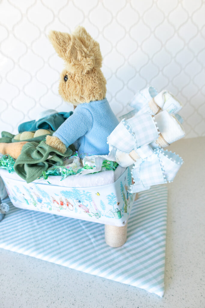 A photo of the back of the Peter Rabbit wheelbarrow diaper cake. A tan plush rabbit with a light blue jacket is sitting in a diaper cake made out of baby blankets and diapers. The handles are made from diapers decorated with a light blue gingham bow. The sides of the wheelbarrow is wrapped in a strip of blue Peter Rabbit fabric. The wheelbarrow tray is propped on cardboard tube legs wrapped with tan yarn.