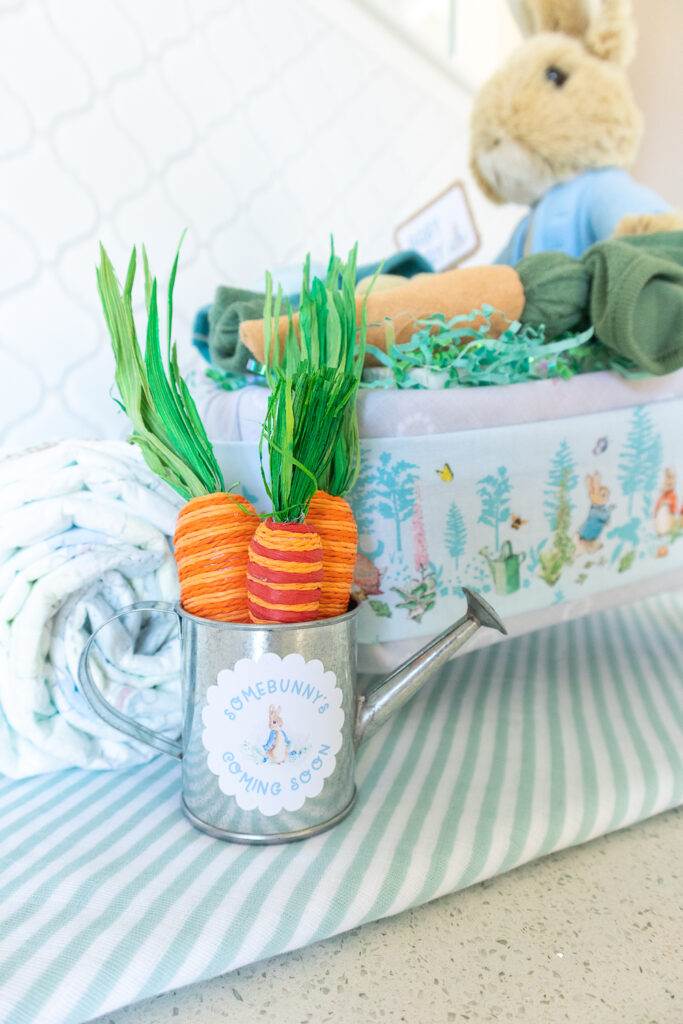 A close up of a mini tin watering can next to the Peter Rabbit wheelbarrow diaper cake. On the can is a sticker that says "Somebunny's coming soon". Three decorative carrots are placed in the watering can. 