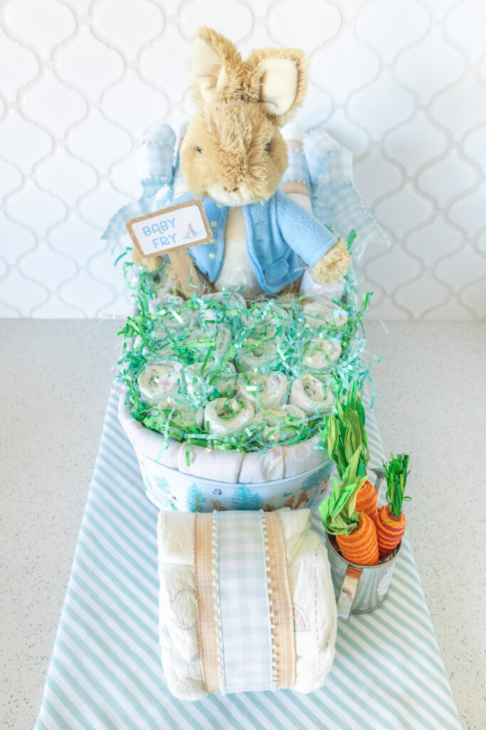 A photo of a Peter Rabbit wheelbarrow diaper cake without the washcloths on top. A tan plush rabbit with a light blue jacket is sitting in a diaper cake made out of baby blankets and diaper. The inside of the wheelbarrow is filled with rolled up diapers and green and aqua easter grass. 