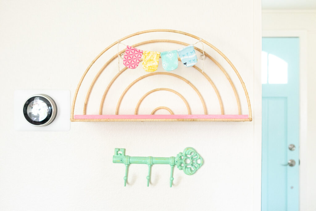 Completed rainbow shaped shelf is hung on a white wall. The wire part of the shelf is wrapped in tan raffia and the wood shelf is painted coral pin. A small handsewn fabric banner is hanging at the top of the shelf. A mint green key shaped hook is hung below the shelf.