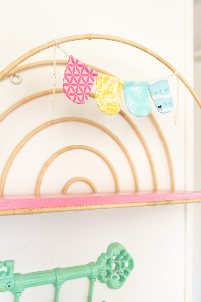 A close-up of the fabric banner on the rainbow wire shelf. There are four hanging fabric scalloped shapes in pink, yellow, mint green and light blue.