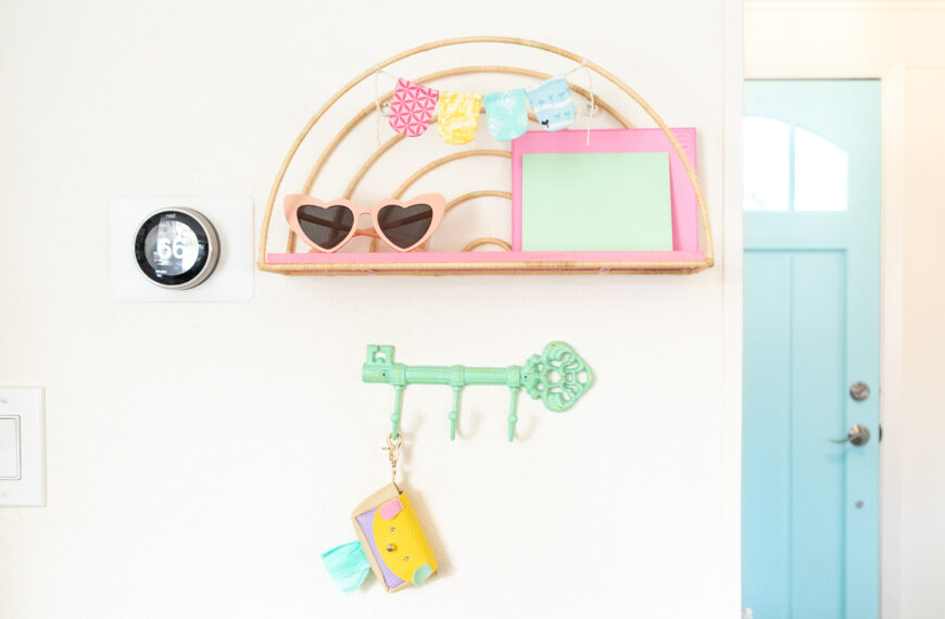 Completed rainbow raffia shelf is hung on a white wall. The wire part of the shelf is wrapped in tan raffia and the wood shelf is painted coral pin. A small handsewn fabric banner is hanging at the top of the shelf. On the shelf is a pair of heart-shaped sunglasses and two colored envelopes. A mint green key shaped hook is hung below the shelf. One of the hooks is holding a pastel puppy pouch made by Blaire.