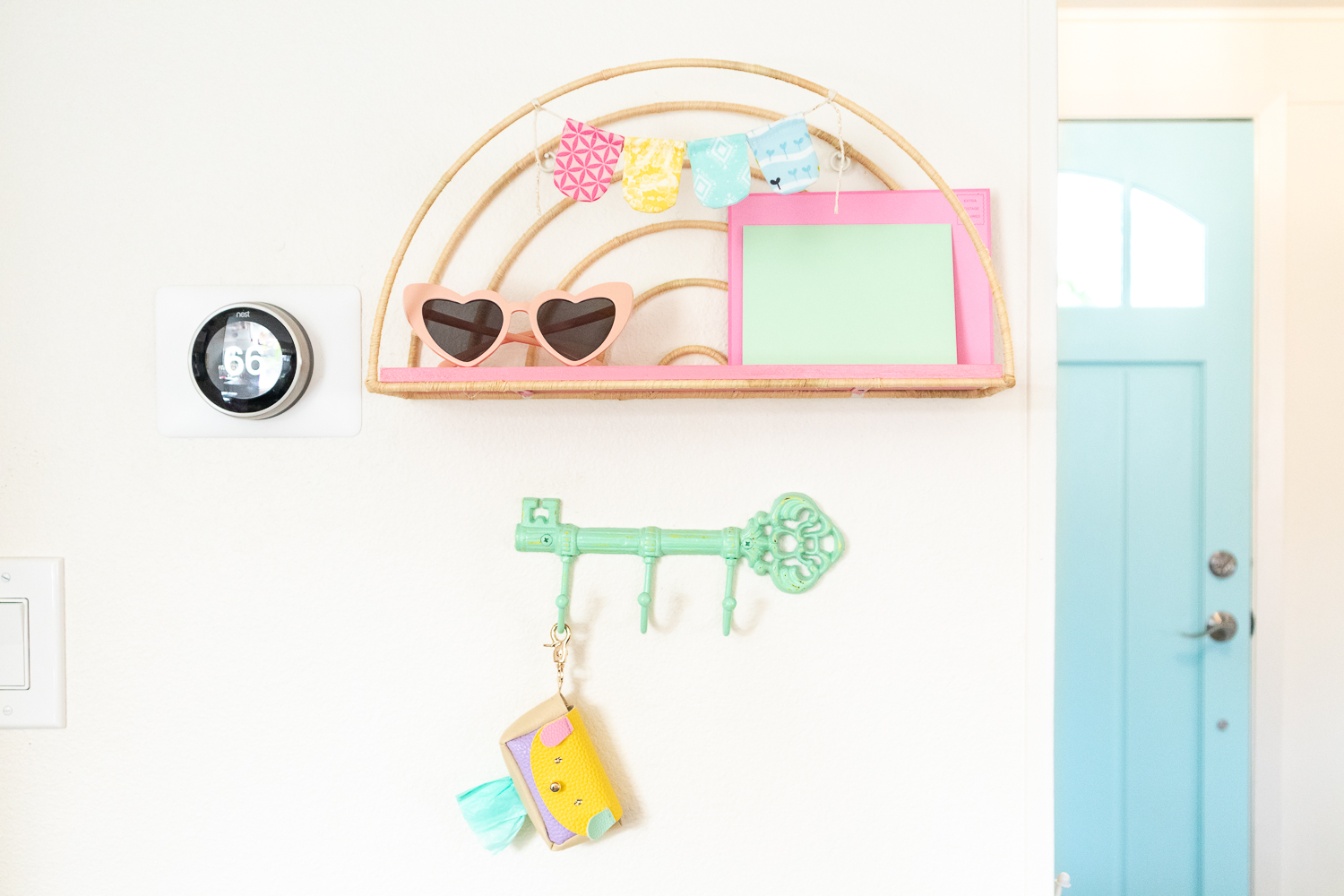 Completed rainbow raffia shelf is hung on a white wall. The wire part of the shelf is wrapped in tan raffia and the wood shelf is painted coral pin. A small handsewn fabric banner is hanging at the top of the shelf. On the shelf is a pair of heart-shaped sunglasses and two colored envelopes. A mint green key shaped hook is hung below the shelf. One of the hooks is holding a pastel puppy pouch made by Blaire.