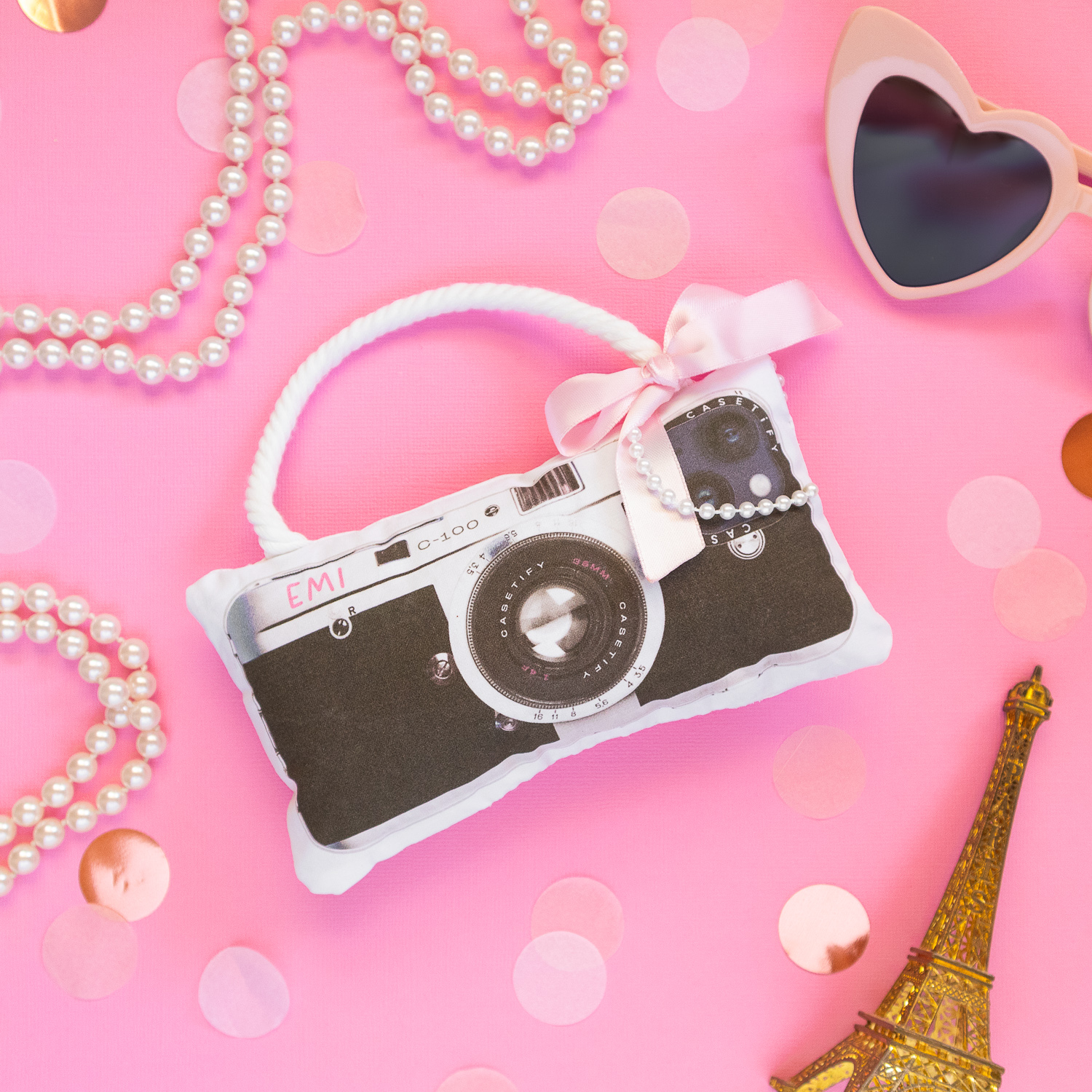DIY Emily in Paris-inspired dog toy made to look like a real vintage camera. The toy is placed on a pink backdrop and is styled with pearls, pink heart sunglasses, an Eiffel tower figuring and light pink confetti.