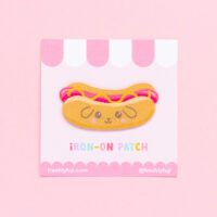 A kawaii hot dog patch with a puppy face on the bun. The woven patch is yellow and pink and is adhered to a cardstock backing with a pink and white striped awning.