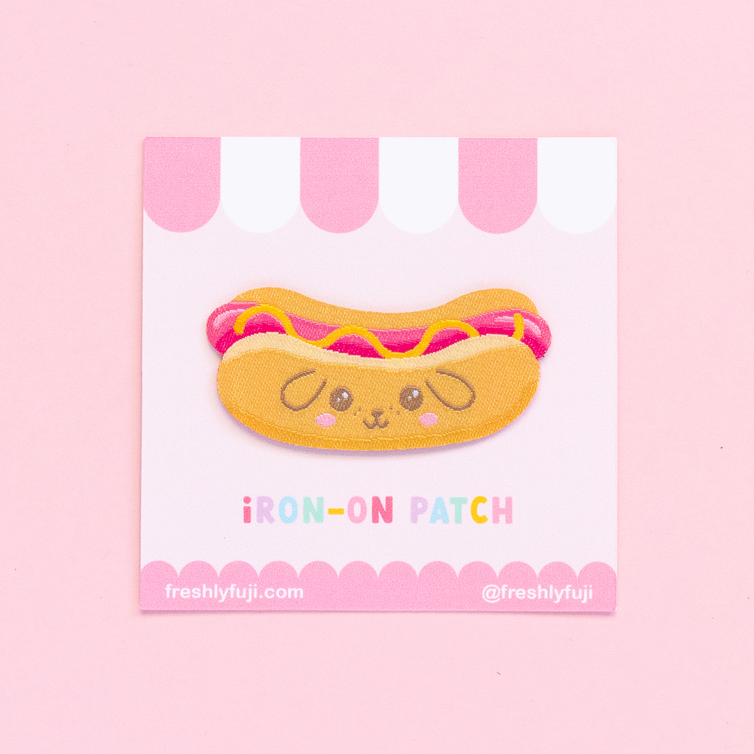 A kawaii hot dog patch with a puppy face on the bun. The woven patch is yellow and pink and is adhered to a cardstock backing with a pink and white striped awning.