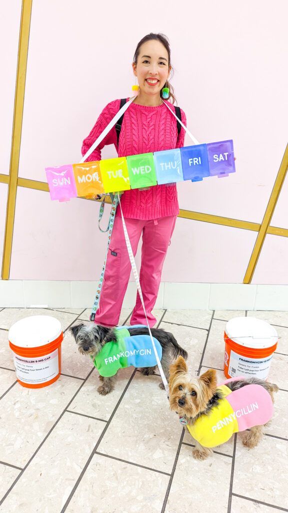 Blaire, a petite Asian American woman, is dressed in hot pink and is wearing a rainbow pillbox costume. Next to her are two small dogs dressed as colorful pills and two orange buckets designed to look like prescription bottles.