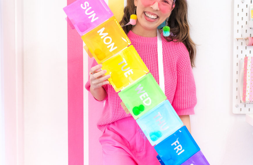 Blaire, an Asian woman with brown hair is wearing a hot pink sweater and pants. She is wearing a rainbow pillbox costume and standing in front of rainbow wall in her craft room.