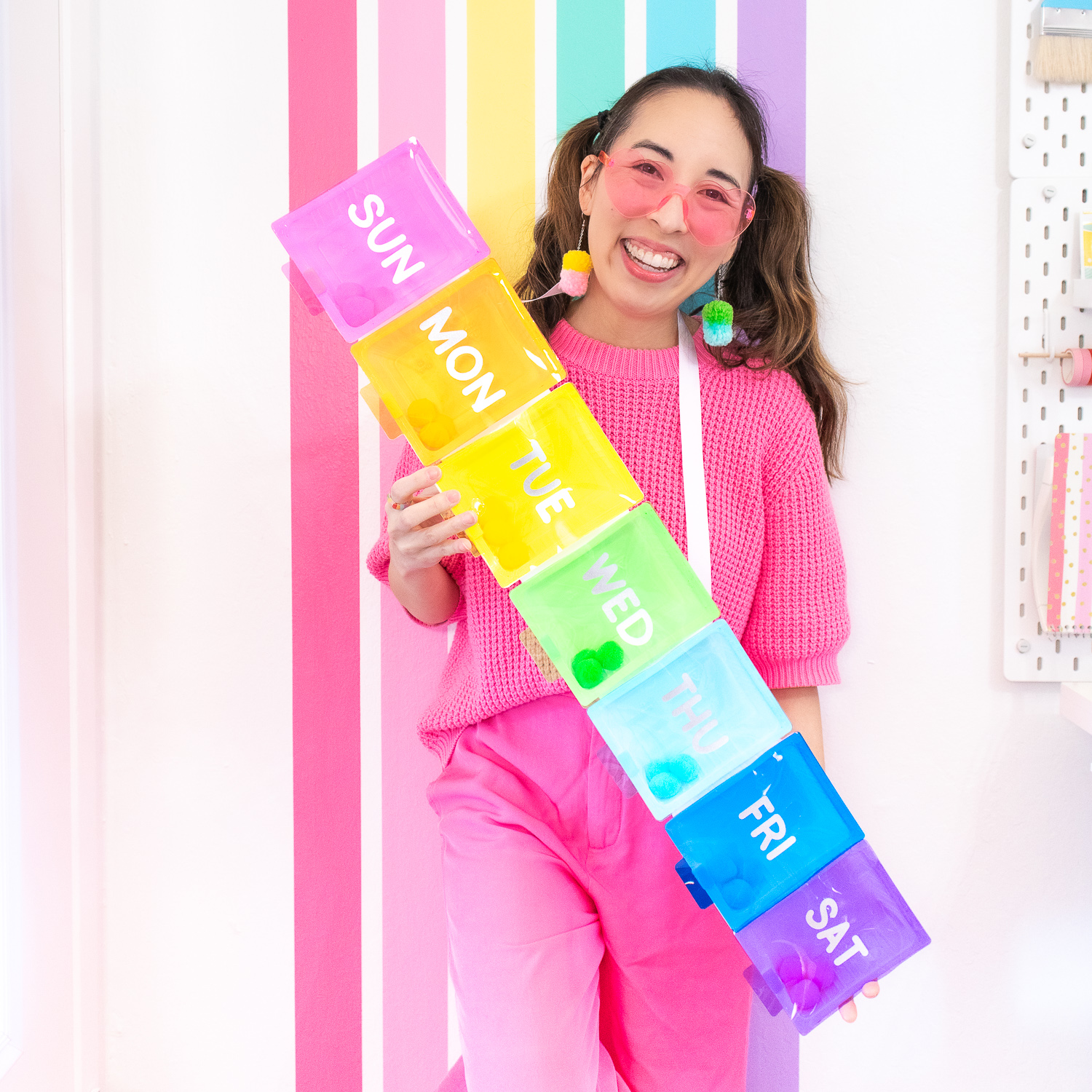 Blaire, an Asian woman with brown hair is wearing a hot pink sweater and pants. She is wearing a rainbow pillbox costume and standing in front of rainbow wall in her craft room.