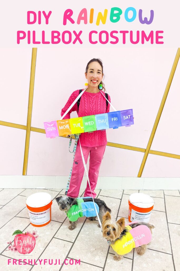 An Asian woman dressed in a hot pink sweater and pants. From her neck hangs a handmade rainbow pillbox costume. At her feet are two small dogs dressed as pills. The gray dog wears a green and blue pill costume with the name "Francomycin". The tan dog wears a yellow and pink pill costume with the name "Pennycillin". There are also two giant orange buckets designed to look like prescription bottles. The top of the photo reads "DIY Rainbow Pillbox Costume".