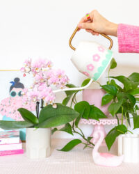 A hand is holding the ivory Ikea Vattenkrase watering can above some flowers and plants. The watering can is decorated with pink flowers and a mint scalloped border made from vinyl.