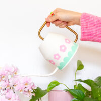 A hand is holding the ivory Ikea Vattenkrase watering can above some flowers and plants. It's decorated with pink flowers and a mint scalloped border made from vinyl.