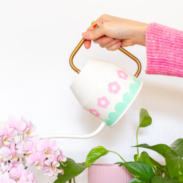 A hand is holding the ivory Ikea Vattenkrase watering can above some flowers and plants. It's decorated with pink flowers and a mint scalloped border made from vinyl.