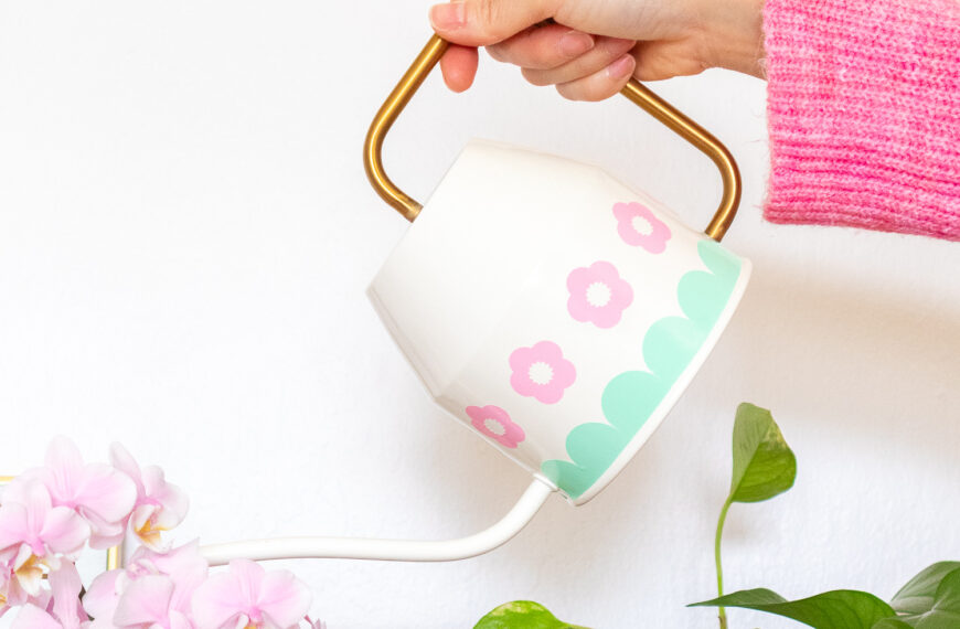 A hand is holding the ivory Ikea Vattenkrase watering can above some flowers and plants. It's decorated with pink flowers and a mint scalloped border made from vinyl.
