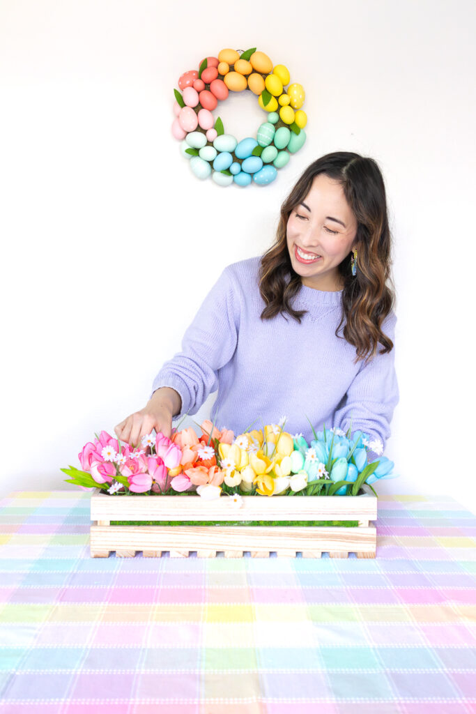 Blaire, an Asian woman with wavy hair is wearing a lavender sweater and is looking down at her pastel rainbow floral centerpiece. It's a wood crate filled with faux tulip flowers in an ombre pattern.