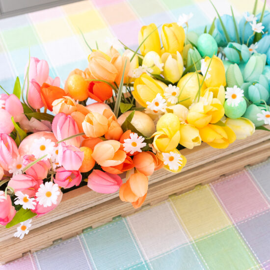 An overhead view of the pastel rainbow floral centerpiece. It's a wood crate filled with faux tulips and capiz eggs arranged in rainbow order. There are small white daisies and mini glitter foam eggs peeking out of the arrangement.