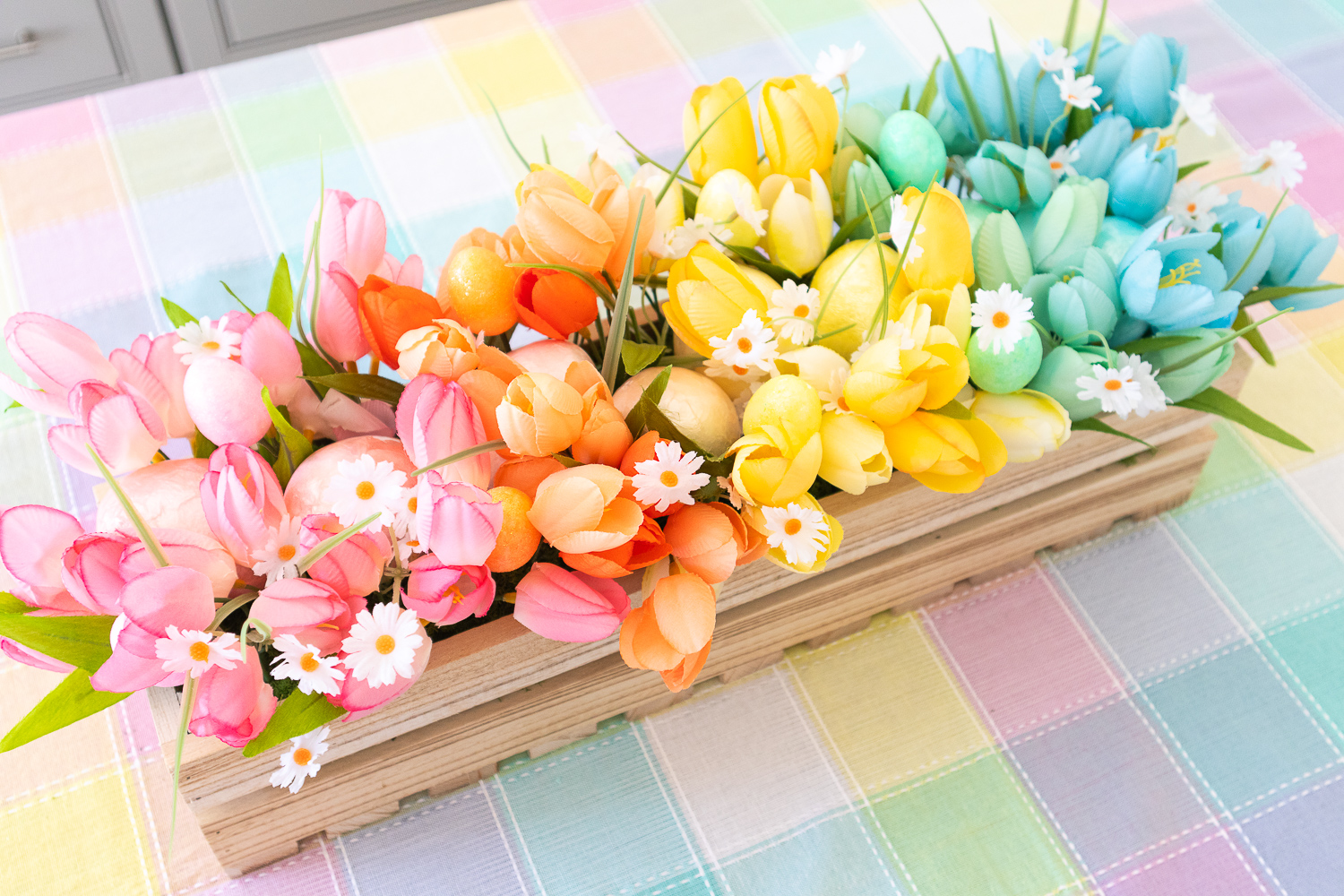 An overhead view of the pastel rainbow floral centerpiece. It's a wood crate filled with faux tulips and capiz eggs arranged in rainbow order. There are small white daisies and mini glitter foam eggs peeking out of the arrangement.