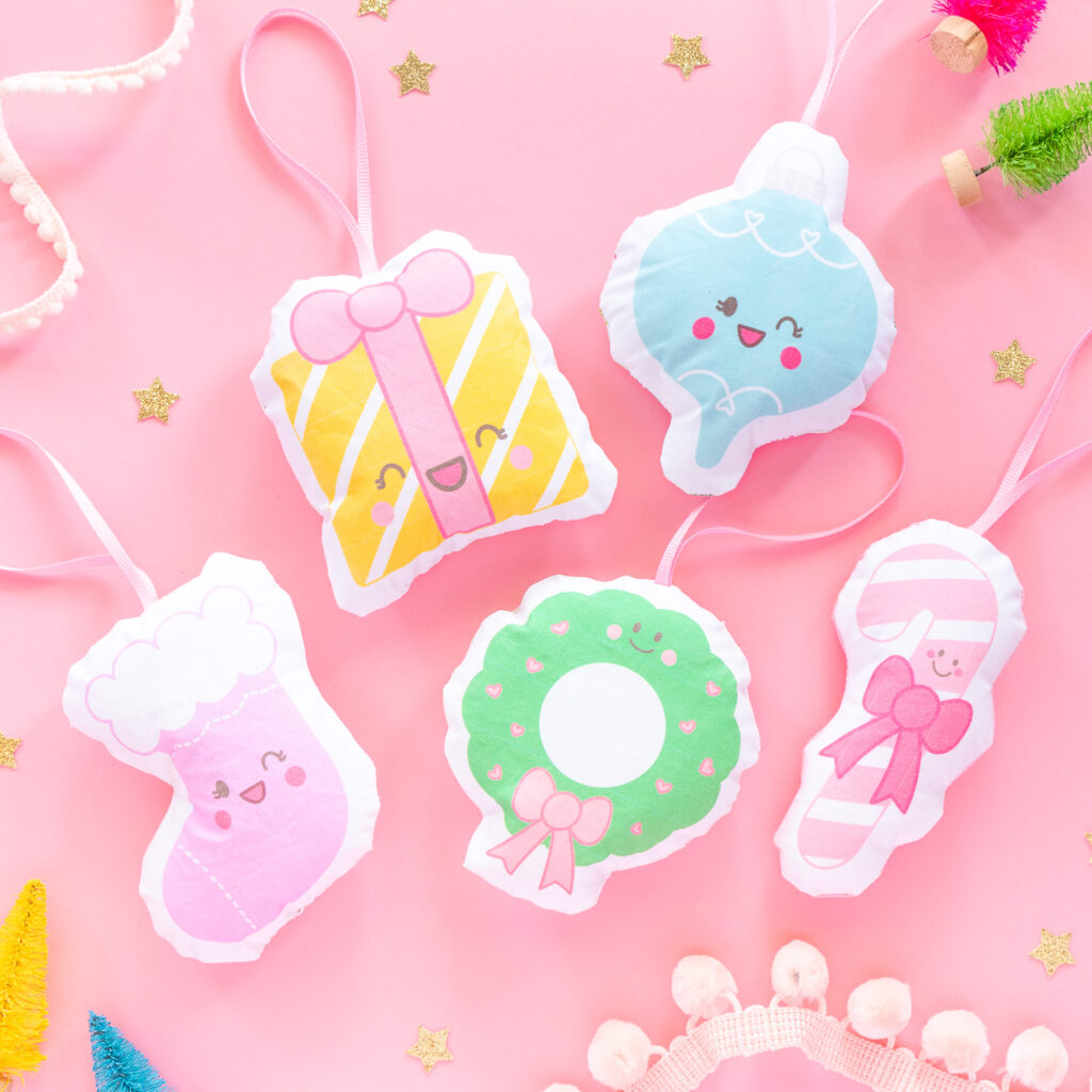 A collection of plush fabric ornaments featuring cute Christmas clipart. There's a stocking, Christmas wreath, candy cane, ornament, and present. The ornaments are scattered on a pink backdrop with tiny bottle brush trees, gold stars, and holiday trim surrounding them.
